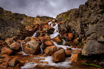 La cascada de agua de las termales naturales desciende por rocas volcánicas, tiñendo el paisaje de tonos ocres y rojizos por el hierro y azufre, bajo un cielo andino dramático.