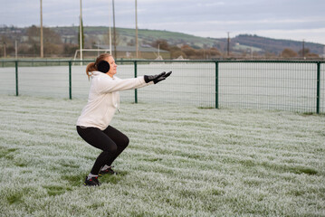 A fit woman in a white hoodie, gloves, and earmuffs performs a squat on a frosty field, outdoor fitness and winter training. Woman doing sports at the stadium, squatting and breathing