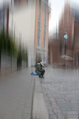 an artistic, atmospheric photograph of a person sitting alone on a wet cobblestone street, surrounded by tall buildings. The vertical motion-blur effect creates a dreamlike, surreal mood
