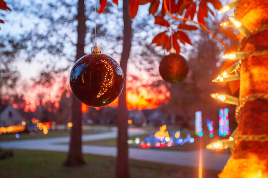 Christmas ornaments hanging from a tree at dusk with glowing holiday lights and a colorful sunset creating a warm, festive neighborhood scene. - Powered by Adobe