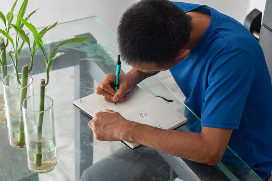 Top view of a man leaning over a glass table writing in a notebook, focused, with visible amputation of his left fingers.