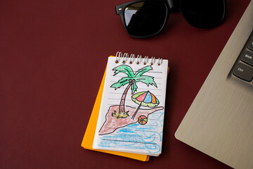 Top view of a small notebook with a hand-drawn sketch of a tropical island beside sunglasses and a laptop on a dark background with copy space, symbolizing work and dream vacation
