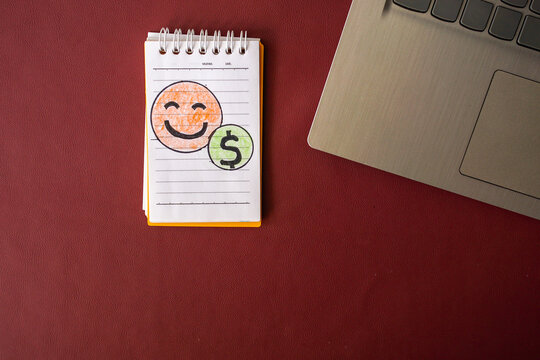 Top view of a small notepad with a hand-drawn smiley face beside a dollar sign and laptop corner on a dark red background with copy space, symbolizing business and financial happiness