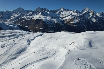 Aerial drone photo of the Swiss Alps showing wide snowy slopes, ski tracks, and dramatic mountain peaks under a clear blue sky. High-altitude winter landscape captured from above.