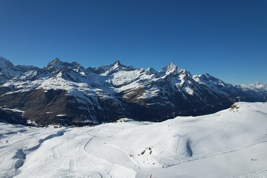 Aerial drone photo of the Swiss Alps showing wide snowy slopes, ski tracks, and dramatic mountain peaks under a clear blue sky. High-altitude winter landscape captured from above. - Powered by Adobe