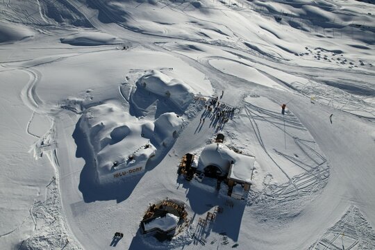 Aerial view of an igloo village surrounded by untouched snow and winter alpine landscapes in the Swiss Alps. Scenic snowy terrain, ski tracks, and unique ice structures under bright winter light.