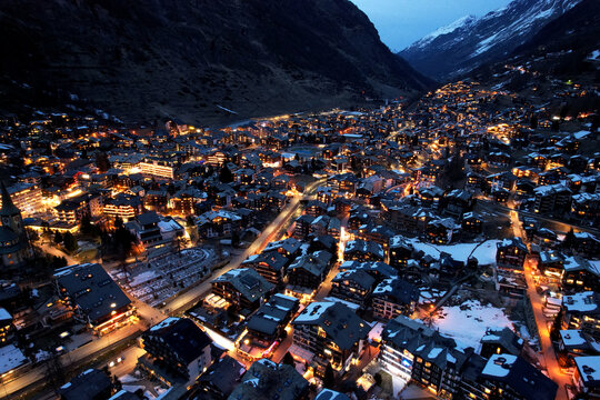 Aerial drone photo of Zermatt at night, showing winter village lights surrounded by snowy mountains and alpine valley scenery.