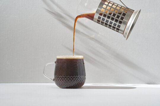 Hot brewed coffee being poured from a metal French press into a textured glass mug in bright natural light, with long soft shadows on a neutral wall creating a clean minimal morning scene
