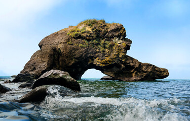 Sea Arch Rock with Waves and Blue Sky. A rugged sea arch rock formation rises from the water under a clear blue sky, with small waves crashing in the foreground.  © kathrineva20
