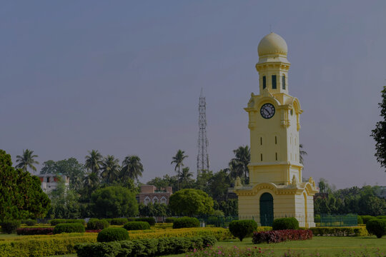 yellow colonial clock tower rises above a landscaped garden under a clear blue sky at hazarduari palace complex, murshidabad