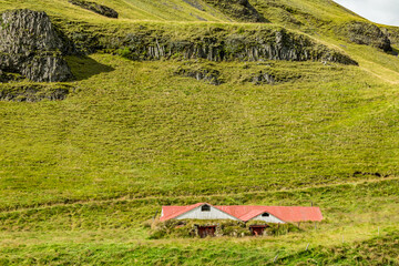Red-roofed turf homes rest quietly beneath a steep green hillside in Southern Iceland&rsquo;s afternoon hush