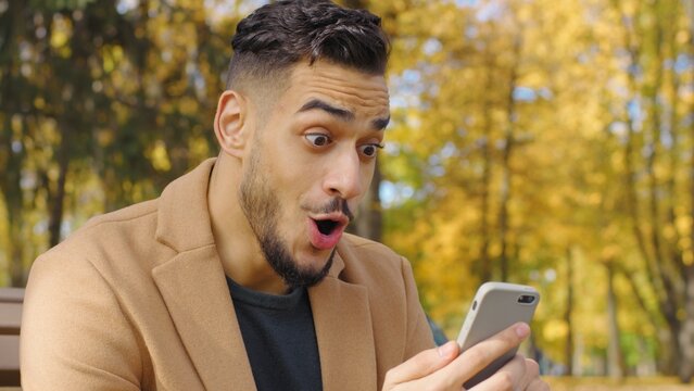 Excited hispanic winner man guy reading good news phone smartphone in autumn park happy male receive message overjoyed internet win victory holding telephone winning success outdoors sitting on bench - Powered by Adobe