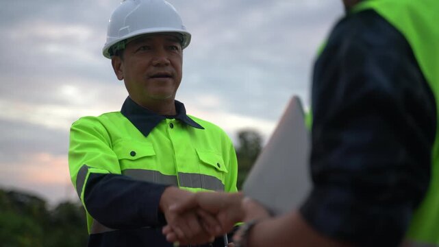Close-up of a confident site manager or engineer in a safety helmet shaking hands with a colleague, symbolizing trust, partnership, and successful project completion on an industrial site at dusk.
