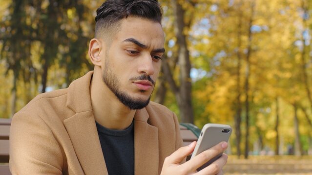 Portrait serious frown Hispanic man using mobile phone guy use app in autumn park reading bad news chat pensive negative emotion relaxing on bench in city looking at smartphone telephone concentrated