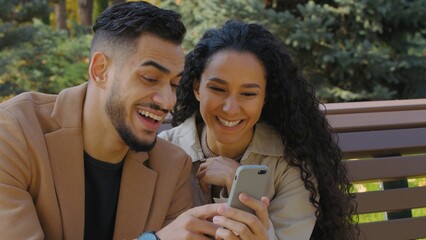 Hispanic happy couple guy with girl in autumn park on bench looking in phone cheerful family man woman discussing shopping online booking restaurant date talking smiling look in mobile app smartphone