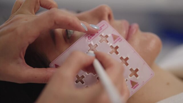 Close-up of unrecognizable beautician preparing female client for facial cosmetic procedure, using skin marking pencil and stencil to outline treatment areas for enhanced beauty, slow motion.