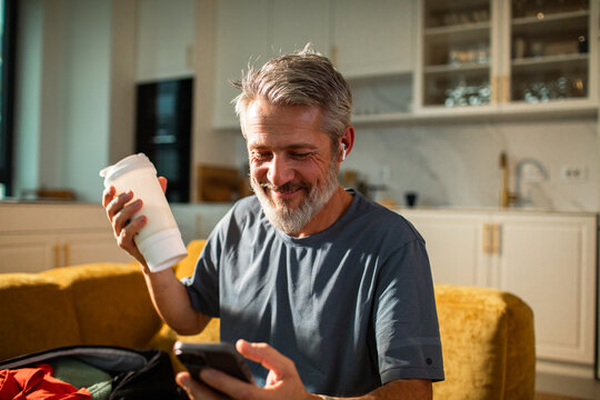 Mature man smiling with a protein shake while using smartphone at home kitchen