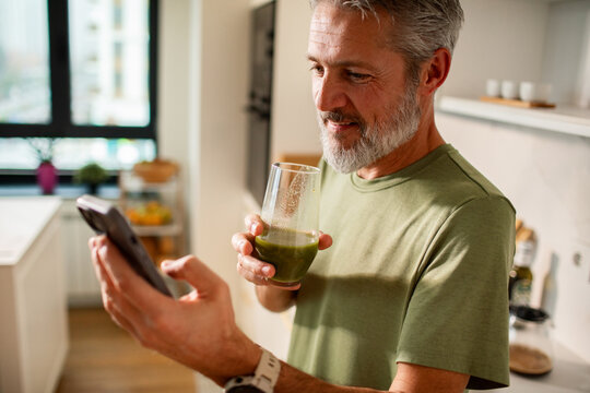 Mature man smiling at phone, drinking green smoothie in home kitchen