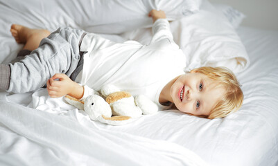 Happy young boy with blond hair lying comfortably in white bedding with a stuffed rabbit toy, smiling at the camera, capturing childhood, comfort, and morning routine