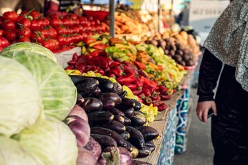 A colorful array of fresh vegetables at a bustling market, featuring tomatoes, peppers, and eggplants, with a shopper in the foreground.