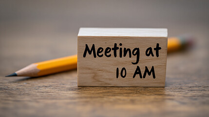 Wooden block with the text "Meeting at 10 AM" placed on a wooden surface beside a yellow pencil, symbolizing organization and time management in a professional setting