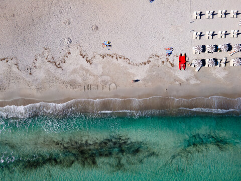 Aerial view of turquoise waters meet the pale sands of Cala Brandinchi, with rows of beach chairs and a bright red lifeguard boat dotting the coastline, San Teodoro, Sardinia, Italy.