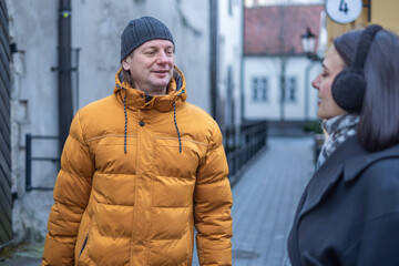 Man wearing orange puffer jacket smiles while talking to a woman in a black coat with ear muffs on a cobblestone street in a charming urban setting with buildings