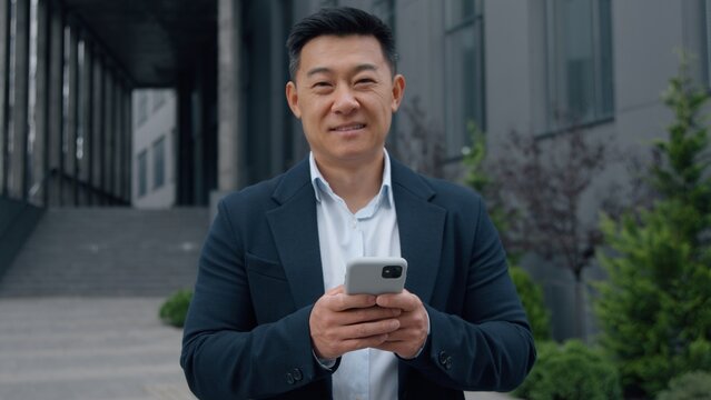 Asian mature businessman holding smartphone Korean business man with mobile phone smiling looking at camera in city happy Japanese male worker Chinese executive smile using application chat internet