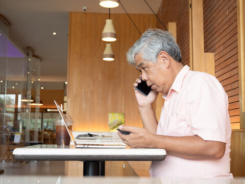 Serious senior Asian man talking on a mobile phone while holding a credit card and using a laptop at a cafe. Active elderly person managing online finance and communications in retirement.