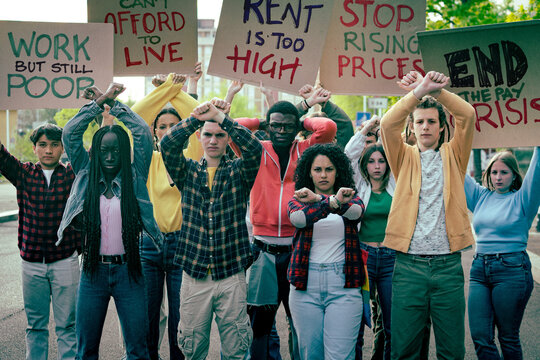 Diverse group of young protesters raising crossed arms and holding signs about rising prices, high rent and low wages during the cost of living crisis, demanding fair pay and affordable living.
