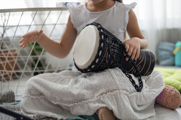 A young girl plays a black drum at home. She wears a white dress and sits on a colorful mat. The...