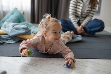 A young Caucasian girl with blonde hair plays on the floor with colorful balls. A woman in a striped shirt sits nearby, creating a cozy home atmosphere.