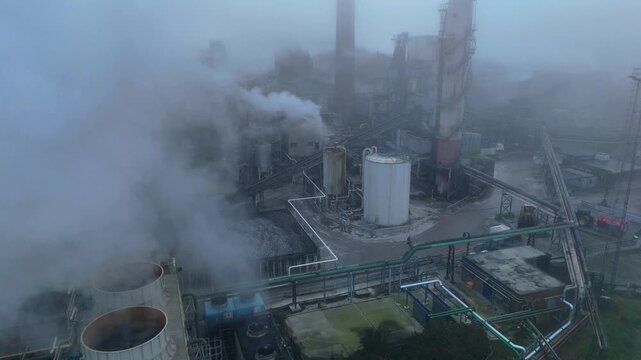 Aerial view of a factory releasing plumes of smoke, its structures partially veiled by fog, creating a stark contrast, Bury Saint Edmunds, England, United Kingdom.