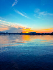 Fototapeta premium A serene sunset over calm waters, with vibrant orange and blue reflections on the surface and a silhouette of a distant bridge and landscape on the horizon.