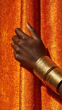 A dark skinned person's hand rests against a shiny orange curtain. The person is wearing two wide, textured gold bracelets and a gold ring