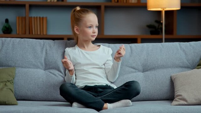 Funny active little Caucasian girl listening music at home dancing clapping fingers dance sitting on couch child kid daughter enjoying musical audio song having fun fooling alone children activity