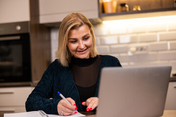 Young woman works on a laptop while taking notes at home in a cozy kitchen setting