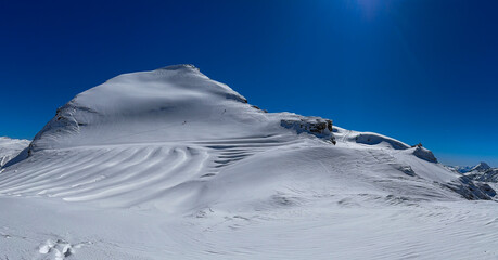 High-altitude snowy ridge with tiny skiers crossing an untouched alpine glacier under a deep blue sky. Serene winter landscape of the Swiss Alps.
