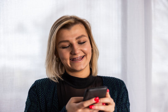 Young woman enjoys texting and smiling while using her smartphone at home during daytime, surrounded by bright light