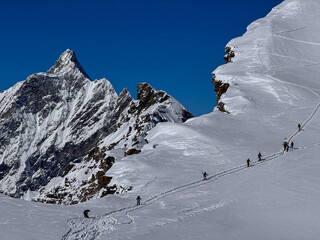 Group of mountaineers climbing a narrow snowy ridge in the Swiss Alps, surrounded by dramatic peaks and deep snow. High-altitude winter adventure and extreme alpine landscape.