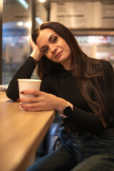 Woman with long dark hair, wearing a black leather jacket, sits at a wooden counter holding a coffee cup, exuding a confident and relaxed vibe in a cozy cafe environment