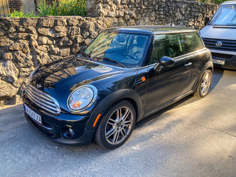 Black Mini Cooper parked on street, near green foliage and stone wall Natural light, angled slightly towards viewer, round headlights, compact shape, no visible tex - Kyiv, Ukraine 07-03-2025