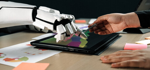 A robotic hand and a human hand engage in collaborative digital art creation on a touchscreen tablet in a modern office setting, showcasing innovation and creativity.Noogenesis
