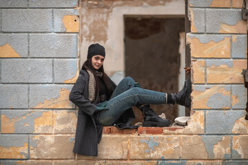 Young woman with long dark hair stands confidently in a broken wall opening, wearing a stylish...