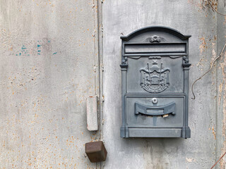 A weathered black mailbox, slightly open with aged decorations, mounted on a wall under subdued lighting Inside is some mail Nearby, a partially visible rusted metal object suggests decay The back