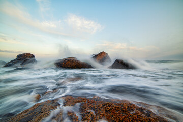 Long-exposure seascape capturing waves swirling around coastal rocks. The soft motion of water and pastel sky create a serene and powerful marine landscape perfect for nature, travel, and relaxation t
