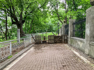 An overcast urban scene featuring a wet brick path leading to an old structure, possibly a bridge or gateway The path is flanked by grass and bare earth, with a rusty metal railing on the left side