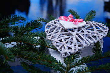 Christmas decorations made of natural materials. On the big heart is a small heart with a red and white ribbon. Against the background - Christmas tree branches and blue water surface.