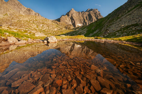 View of jagged peaks mirroring in the still, clear waters revealing rocky depths, framed by grassy slopes under a bright sky, Tatranska Javorina, Presovsky kraj, Slovakia.
