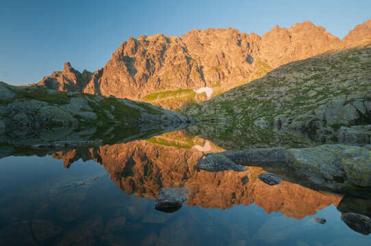 View of golden sunlight kissing the jagged peaks reflected in the still waters of Hrube pleso tarn, creating a serene mountainscape, Tatranska Javorina, Presovsky kraj, Slovakia.